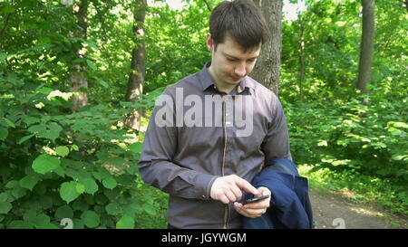 Junger Mann im Wald steht, denkt an etwas und funktioniert auf dem Handy. Stockfoto