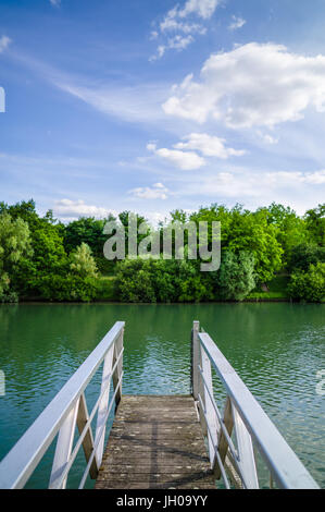 Ein Holzponton mit metallischer Handlauf über einen Fluss mit Bäumen und Vegetation unter blauem Himmel mit weißen Wolken. Stockfoto