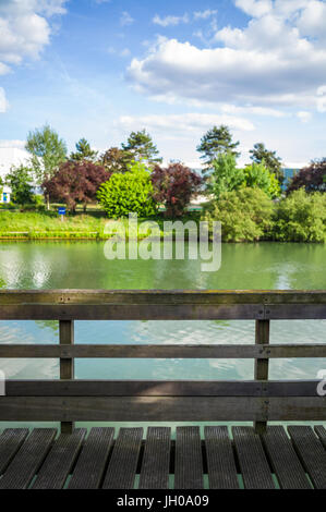 Ein Holzponton mit Handlauf aus Holz vor einem Fluss mit Bäumen und Vegetation unter blauem Himmel mit weißen Wolken. Stockfoto