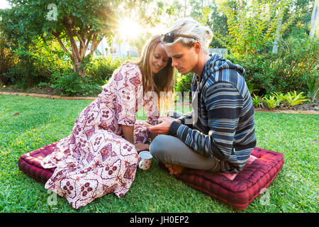 Ein junges Paar mit Blick auf ein Telefon in einem lebendigen Garten als helle Sonne sitzend Licht scheint durch die Bäume. Stockfoto