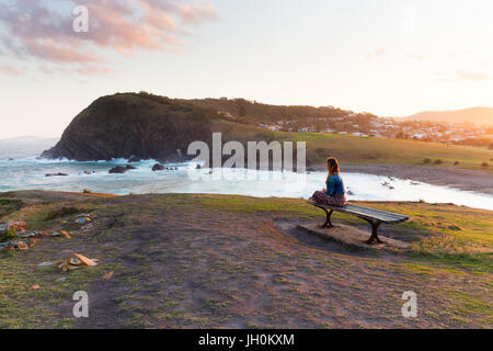 Eine Frau sitzt auf einer Holzbank und Uhren die goldene Sonnenlicht verschwinden aus der wunderschönen Küste um sie herum.