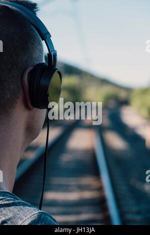 Nahaufnahme eines jungen Mannes gesehen von hinten mit Kopfhörern neben den Bahngleisen Stockfoto