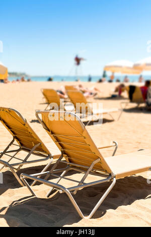 Nahaufnahme von einigen beigen Sonnenliegen und einige Beige Schirme in einem Strand im Mittelmeer, mit vielen unkenntlich Menschen Sonnenbaden in den Rücken Stockfoto