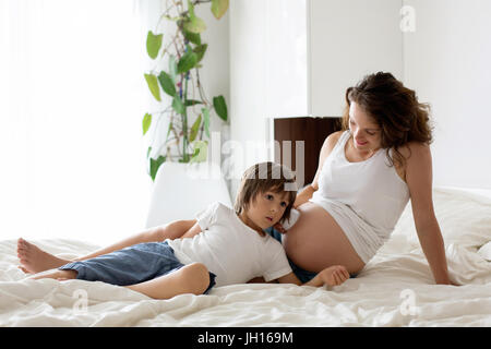 Schöne schwangere junge Mutter, sitzen im Bett mit ihrem älteren Kind, für das ungeborene Kind durch den Bauch hören Stockfoto