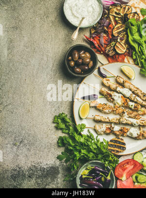 Sommer Grill Party Dinnerset. Flatlay von gegrilltem Hähnchen-Spieße mit Joghurt-Dip, Fladenbrot, Petersilie, Gemüse, marinierten Oliven, Chilis über gr Stockfoto