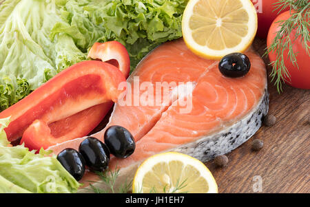 Roher Lachs Steak auf einem Holzbrett. Blätter für Salat und Dill, ein Tomaten-Zweig, schwarze Oliven, Gewürze, Scheiben Zitrone und Paprika, Erbsen Kuvertierung Stockfoto