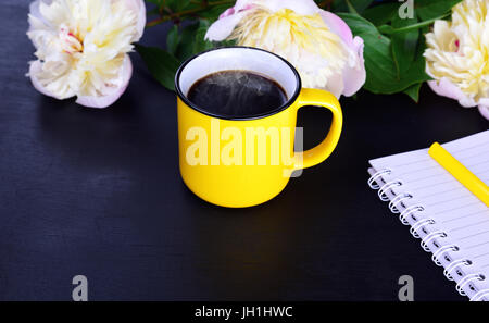 Heißen schwarzen Kaffee in einem gelben Becher auf schwarzem Hintergrund aus Holz mit einem Bouquet von weiß blühende Pfingstrosen Stockfoto