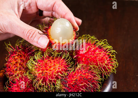 Nahaufnahme einer Frau Hand hält eine Rambutan, die offen in der Nähe von einer Schüssel mehr diese köstliche Frucht geschnitten worden Stockfoto