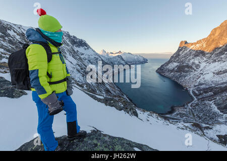 Wanderer auf dem Kamm bewundert das Dorf Fjordgard umrahmt das gefrorene Meer des Ornfjorden, Senja, Troms, Norwegen Stockfoto