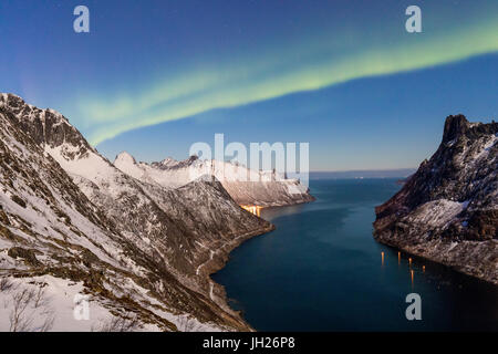 Northern Lights auf die schneebedeckten Gipfel und das Dorf Fjordgard umrahmt das gefrorene Meer, Ornfjorden, Senja, Troms, Norwegen Stockfoto