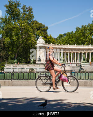 Frau mit dem Fahrrad in Madrids Park del Retiro / Parque del Retiro vorbei an den See mit dem Denkmal König Alfonso XII n der Rückseite geschliffen Stockfoto