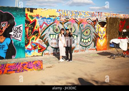 Ein Ehepaar aus Hongkong filmt selbst mit einer kleinen Kamera, wie sie vor Graffiti auf einem verbliebenen Teil der ehemaligen Berliner Mauer am Mauerpark (Wand Parc) in Berlin am 18. Juni 2017 stehen. Dieses Bild ist Teil einer Serie von Fotos auf den Tourismus in sein | weltweite Nutzung Stockfoto
