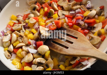 Overhead Schuss von Pilzen, roten Zwiebeln und rote und gelbe Paprika mit alten hölzernen Spatel in einer keramischen Pfanne braten. Stockfoto