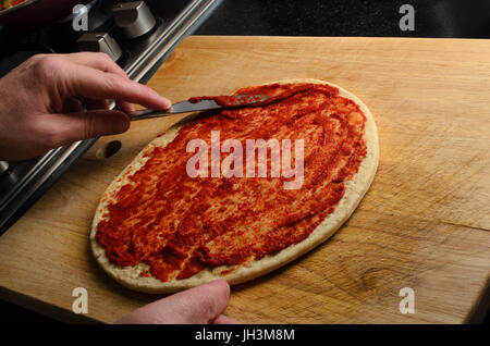Männliche übergeben Pizzaboden mit Stahl Messer auf verkratzt und abgenutzt Schneidebrett Verbreitung Tomatenmark oder Sauce.  Schwarze Oberfläche und Rand der Koch Stockfoto