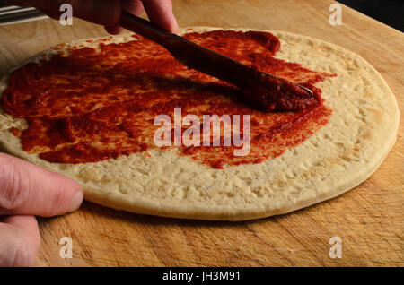 Männliche Hand Tomaten-Basis über Pizza auf abgenutzte, zerkratzte Schneidbrett aus Holz verbreitet.  Dunkle Beleuchtung. Stockfoto