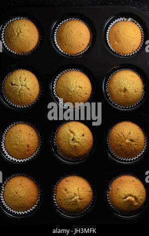 Aufwand für ein Dutzend goldene Tasse Kuchen gerade aus dem Ofen gebacken in Papier Fällen und noch innen schwarz Brötchen Zinn. Stockfoto