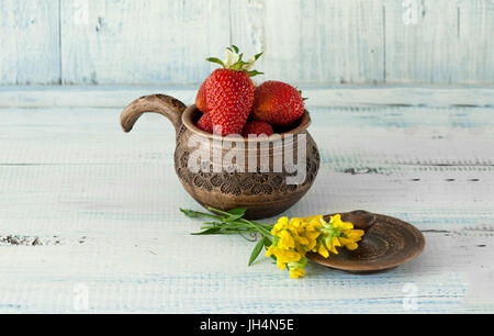 Erdbeeren rot, reif liegt in einem braunen Tonkrug mit Griff mit kleinen gelben Blüten auf blauem Grund aus Holz. Die horizontalen Rahmen. Stockfoto