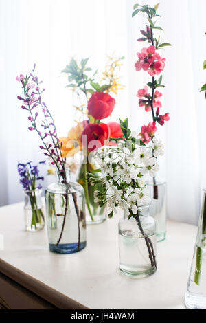 Verschiedene schöne Blumen in Gläser mit Wasser auf dem Tisch neben dem Fenster Stockfoto