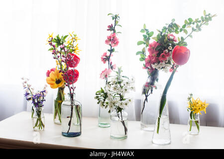 Verschiedene schöne Blumen in Gläser mit Wasser auf dem Tisch neben dem Fenster Stockfoto