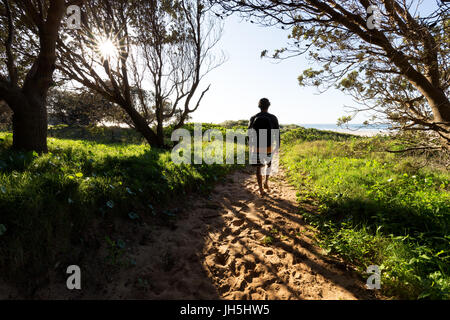 Ein Mensch lebt auf einem sandigen Pfad durch Bäume und hinterleuchtete, beleuchteten Rasen in Richtung Strand. Stockfoto