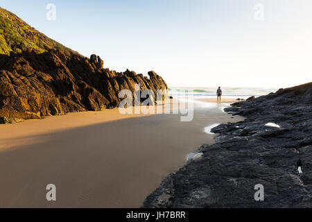 Eine Person steht allein an einem einsamen Strand in Australien und sieht die helle Morgensonne leuchten die Küste und das Meer, die Sie umgibt. Stockfoto