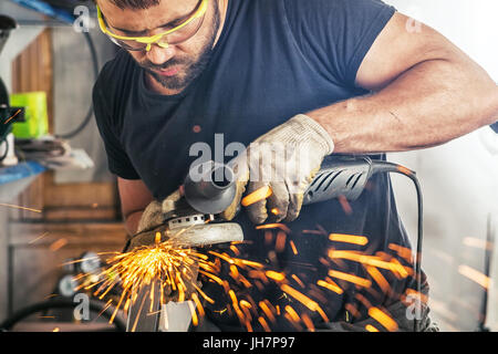 Ein junger Mann, der Schweißer auf ein schwarzes T-Shirt, Schutzbrille und Handschuhe Prozesse Metall ein Winkelschleifer in der Garage, im Hintergrund eine Menge zu Stockfoto