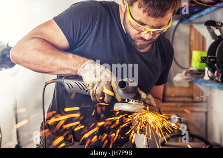 Ein junger Mann, der Schweißer auf ein schwarzes T-Shirt, Schutzbrille und Handschuhe Prozesse Metall ein Winkelschleifer in der Garage, im Hintergrund eine Menge zu Stockfoto