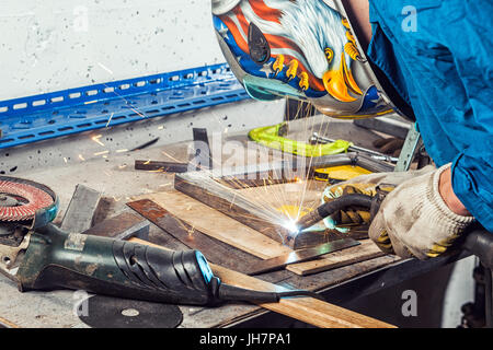 Ein Mann Schweißer auf einer blauen insgesamt, das Tragen eines Schweißen Maske mit einem Adler und Bau Handschuhe, schweißen Sie ein Metall Schweißen Maschine in einer Werkstatt, Funken fliegen, Stockfoto