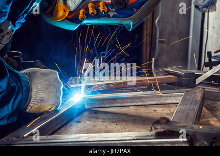 Ein junger Mann, der Schweißer auf einer blauen Bau die in einem schweissverfahren maske und handschuhe, schweißen Sie ein Metall Produkt mit einem Schweißgerät in den Werken Stockfoto