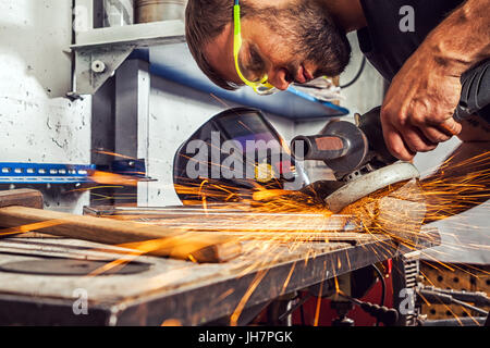 Ein junger Mann, der Schweißer eine Brünette in einem schwarzen T-Shirt und Schutzbrille. Er verarbeitet eine Metall Element aus einem Winkelschleifer in der Werkstatt. close-up auf der s Stockfoto