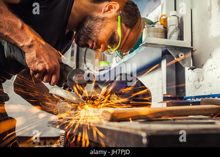 Ein junger Mann, der Schweißer eine Brünette in einem schwarzen T-Shirt und Schutzbrille. Er verarbeitet eine Metall Element aus einem Winkelschleifer in der Werkstatt. close-up auf der s Stockfoto