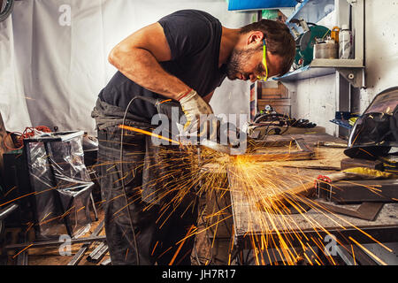 Eine junge Brünette mann Schweißer in einem schwarzen T-Shirt, Schutzbrille, Prozesse ein Metall Produkt mit einem Winkel gringer in der Garage, seitwärts Funken Stockfoto