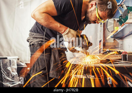 Eine junge Brünette mann Schweißer in einem schwarzen T-Shirt, Schutzbrille, Prozesse ein Metall Produkt mit einem Winkelschleifer in der Garage, seitwärts Funken Stockfoto