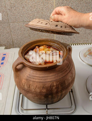 Sauer gefüllte Kohlblätter mit Speck auf der Oberseite im traditionellen Tontopf. Eine alte weibliche Hand hält den Deckel. Der Topf wird auf dem Herd. Stockfoto