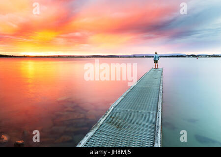 Eine Person Uhren einen schönen, farbenfrohen Sonnenuntergang über dem Meer aus dem Ende des einen kleinen Steg. Stockfoto