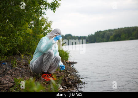 Foto des Chemikers im medizinischen Anzug und Maske nehmen Wasserproben im Teich Stockfoto