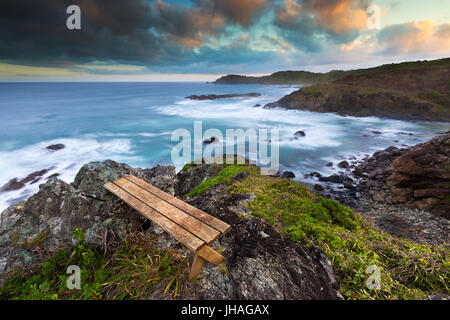 Eine leere Holzbank mit Blick auf eine schöne Felsküste bei Sonnenuntergang in Australien. Stockfoto