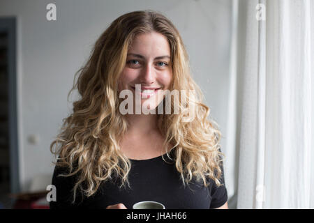 Jung, blond, schöne kaukasischen Frau bei einem heißen Getränk in der Nähe der Fenster im natürlichen Morgenlicht. Drinnen im Sommer auf einer Canon 5D aufgenommen Stockfoto