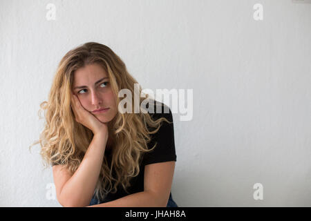 Jung, blond, Kaukasische Frau verärgert beim hinsetzen. Auf der Suche nach außerhalb des Bildschirms und nicht Blickkontakt vor einem weißen Hintergrund in natürlichem Licht. Stockfoto