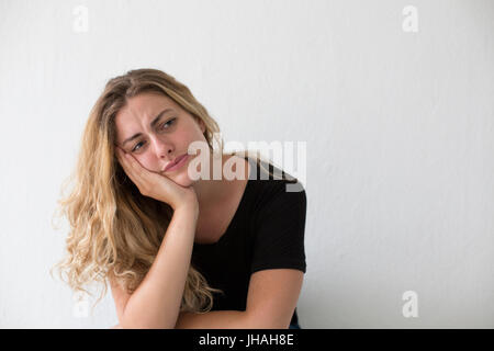 Jung, blond, Kaukasische Frau verärgert beim hinsetzen. Auf der Suche nach außerhalb des Bildschirms und nicht Blickkontakt vor einem weißen Hintergrund in natürlichem Licht. Stockfoto