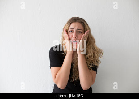 Jung, blond, schön, Milennial Frau mit dem Ausdruck ihrer Aufregung und Verwirrung vor einem weißen Hintergrund und in einem schwarzen Shirt im Haus während des Tages. Stockfoto