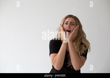 Jung, blond, schön, Milennial Frau mit dem Ausdruck ihrer Aufregung und Verwirrung vor einem weißen Hintergrund und in einem schwarzen Shirt im Haus während des Tages. Stockfoto