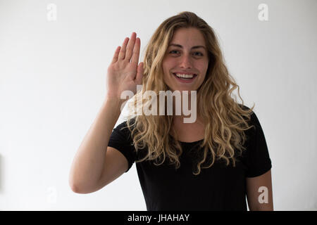 Jung, blond, kaukasischen Milennial Frau Gefühle, Freude auszudrücken und während ihre Hand auf den Betrachter vor einem weißen Hintergrund winken lächelnd. Stockfoto