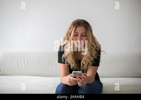 Jung, blond, schön Milennial Frau Glück während des Spiels auf ihrem Smartphone, sitzen auf der Couch vor einem weißen Hintergrund zum Ausdruck zu bringen. Stockfoto