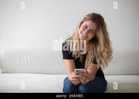 Jung, blond, schön Milennial Frau Glück während des Spiels auf ihrem Smartphone, sitzen auf der Couch vor einem weißen Hintergrund zum Ausdruck zu bringen. Stockfoto