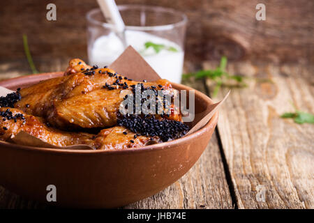 Würzige Hähnchenflügel mit Sesam in Keramikschale serviert mit Joghurt-Dip auf rustikalen Holztisch glasiert Stockfoto