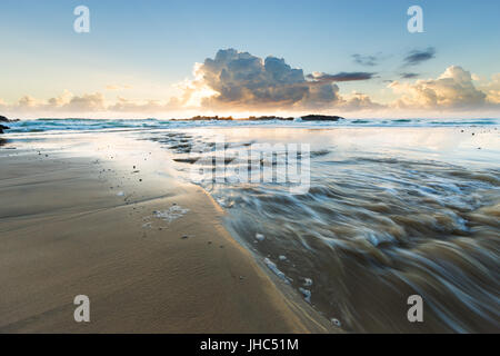 Ein frisches Wasser fließt ins Meer am leeren Strand in Australien wie Gewitterwolken durch die Morgensonne beleuchtet sind. Stockfoto