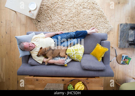 Ältere Mann liegt auf dem Sofa im Wohnzimmer seines Hauses. Er hat seinen Hund lag auf ihm, der auch im Ruhezustand befindet. Stockfoto