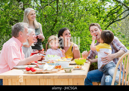 Drei-Generationen-Familie mit einer Gartenparty im Sommer. Jeder lacht und schaut der kleine Junge, der seinen Vater einige rote Pe Fütterung ist Stockfoto