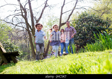 Ein kleiner Junge und seine Schwester laufen in den Rasen in einem Naturpark. Ihre Eltern sind unscharf im Hintergrund, hinter ihnen gehen. Stockfoto